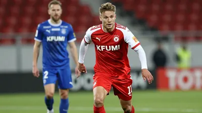 A tight action crop of a KFUM Oslo player in a red jersey with white sleeves, running with the ball, with a blurred...